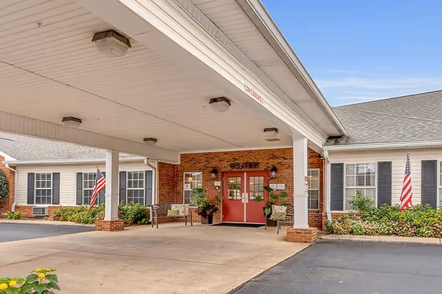 Entrance of a senior living facility with a covered drop-off area supported by white columns. The building has red double doors with the word 'Welcome' above them, brick walls, white siding, black shutters on windows, and American flags on either side of the entrance. There are plants and benches near the entrance.