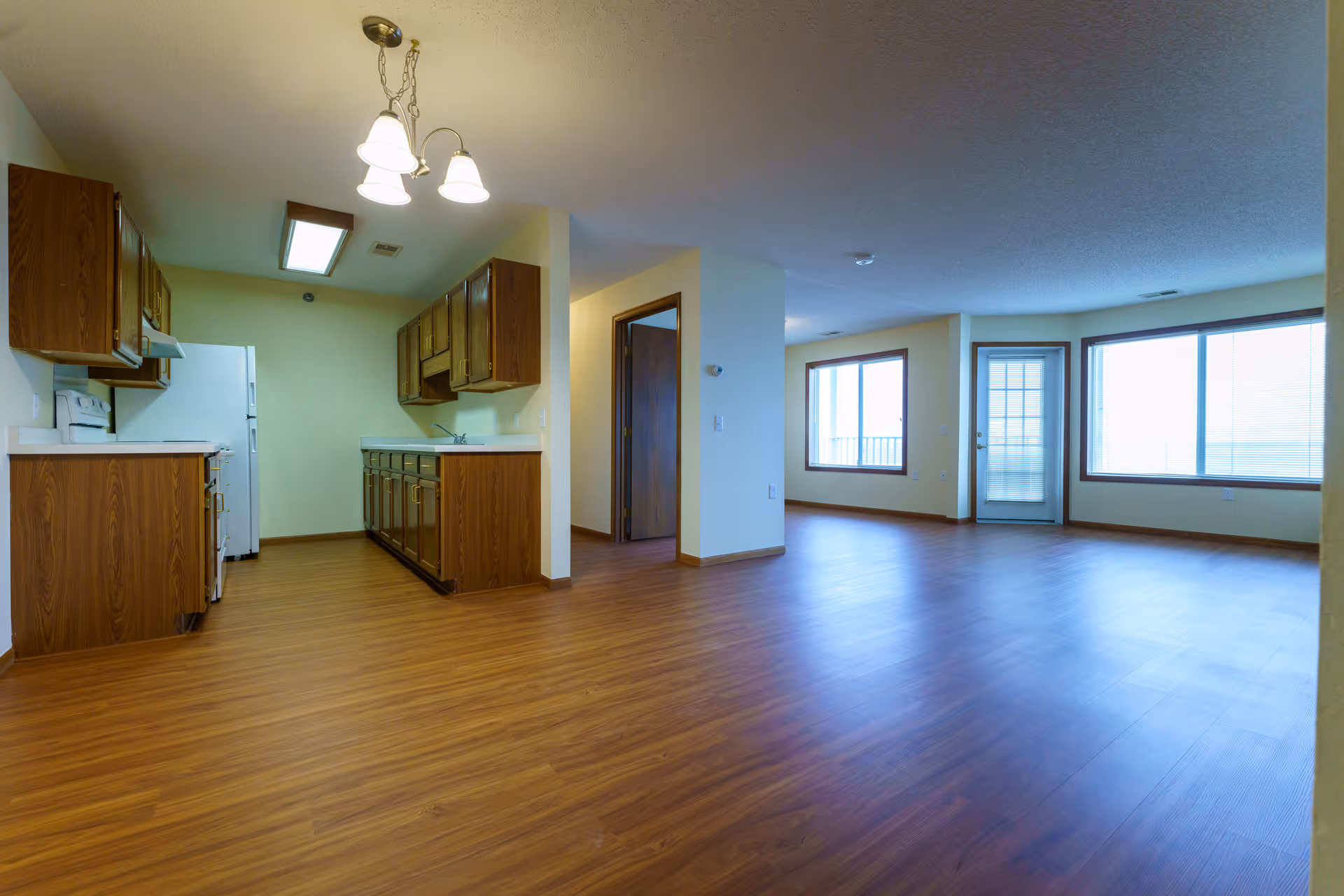 Interior view of an empty apartment featuring a kitchen with wooden cabinets and white appliances on the left, and a spacious living area with large windows and a glass door leading outside on the right. The floor is covered with wood-like laminate flooring and the walls are painted light yellow.