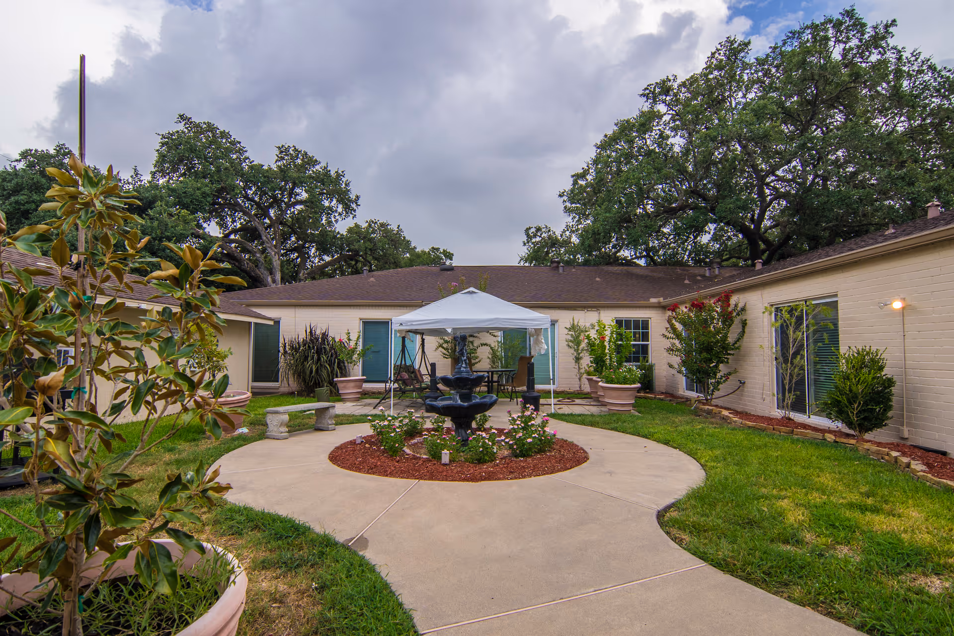 Outdoor courtyard area of Richmond Health Care Center with a circular concrete pathway surrounding a central flower bed with a black fountain. There is a white canopy tent with chairs underneath, large potted plants, and trees surrounding the courtyard. The building walls are light-colored with several windows and doors.