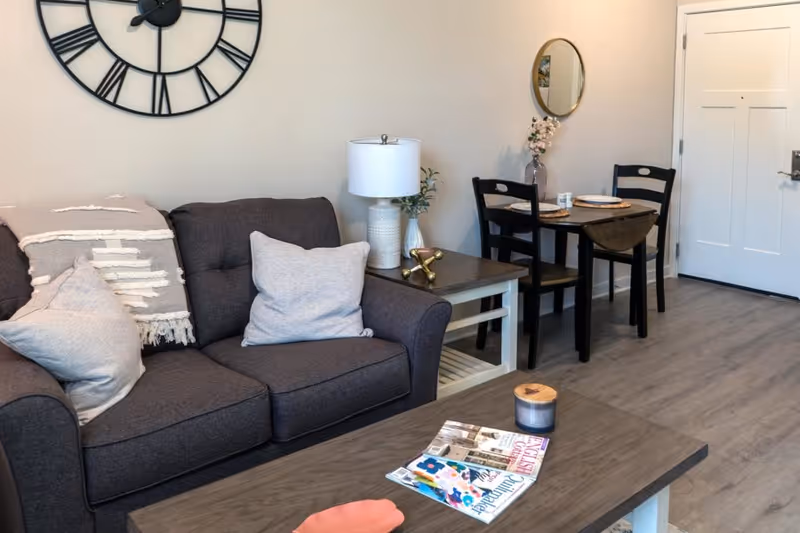 Cozy living room with a gray sofa, coffee table with magazines and a candle, a large wall clock, and a small dining table with two chairs by the entry door.
