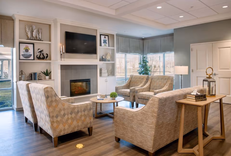 A cozy living room area with four patterned armchairs and a beige sofa arranged around a round wooden coffee table. The room features a modern electric fireplace with a mounted flat-screen TV above it, built-in shelves with decorative items, large windows with blinds, a floor lamp, and a wooden side table with books and a lantern.