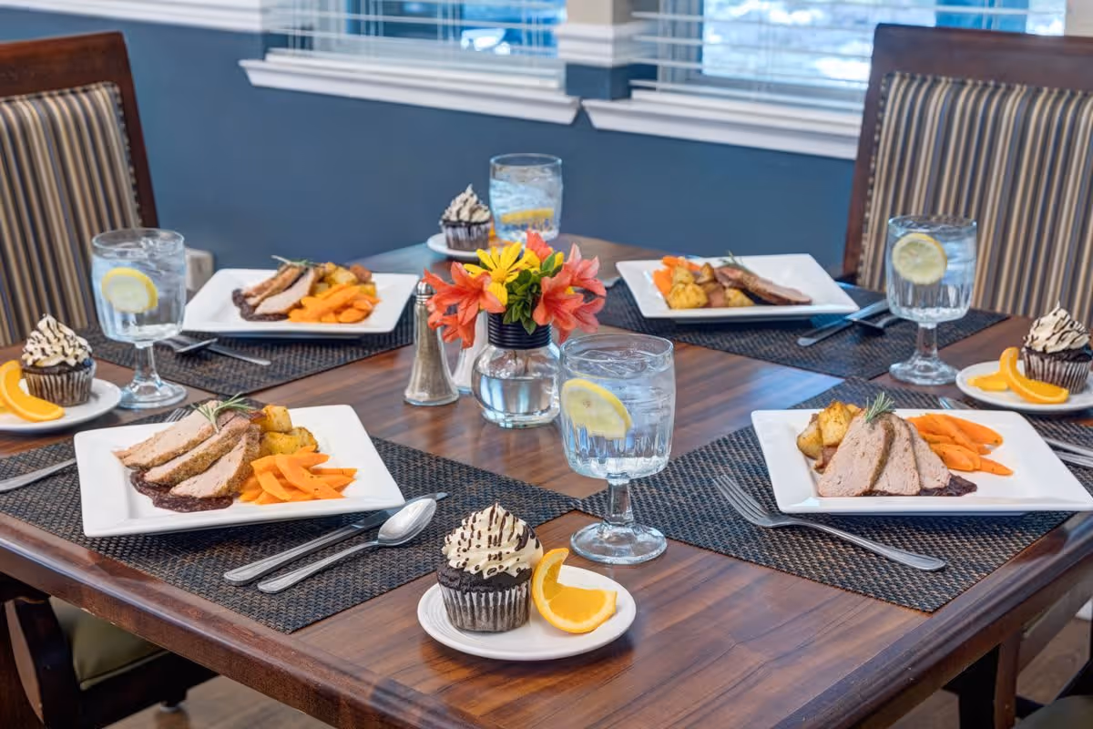 A dining table set for four with plates of sliced meat, roasted potatoes, and cooked carrots. Each place setting includes a glass of water with a lemon slice, silverware, and a small plate with a chocolate cupcake topped with cream and a slice of orange. A small vase with colorful flowers is in the center of the table.