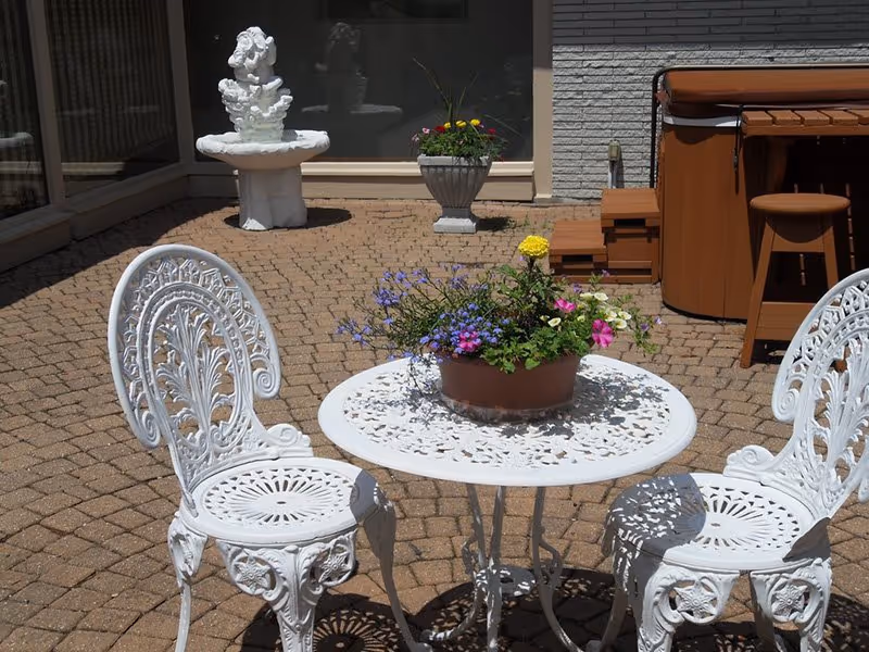 White ornate metal patio table and chairs on a brick-paved courtyard with a potted flower centerpiece, fountain, and hot tub in the background.