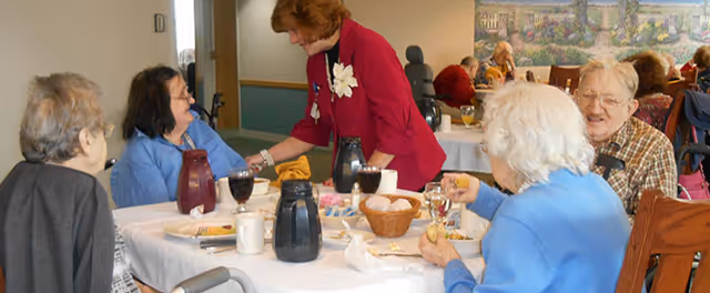 Elderly residents seated around a dining table in a communal dining room while a staff member assists them.