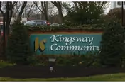 Outdoor view of a green sign with the text 'Kingsway Community' surrounded by bushes and trees, with a parked car and building partially visible in the background.