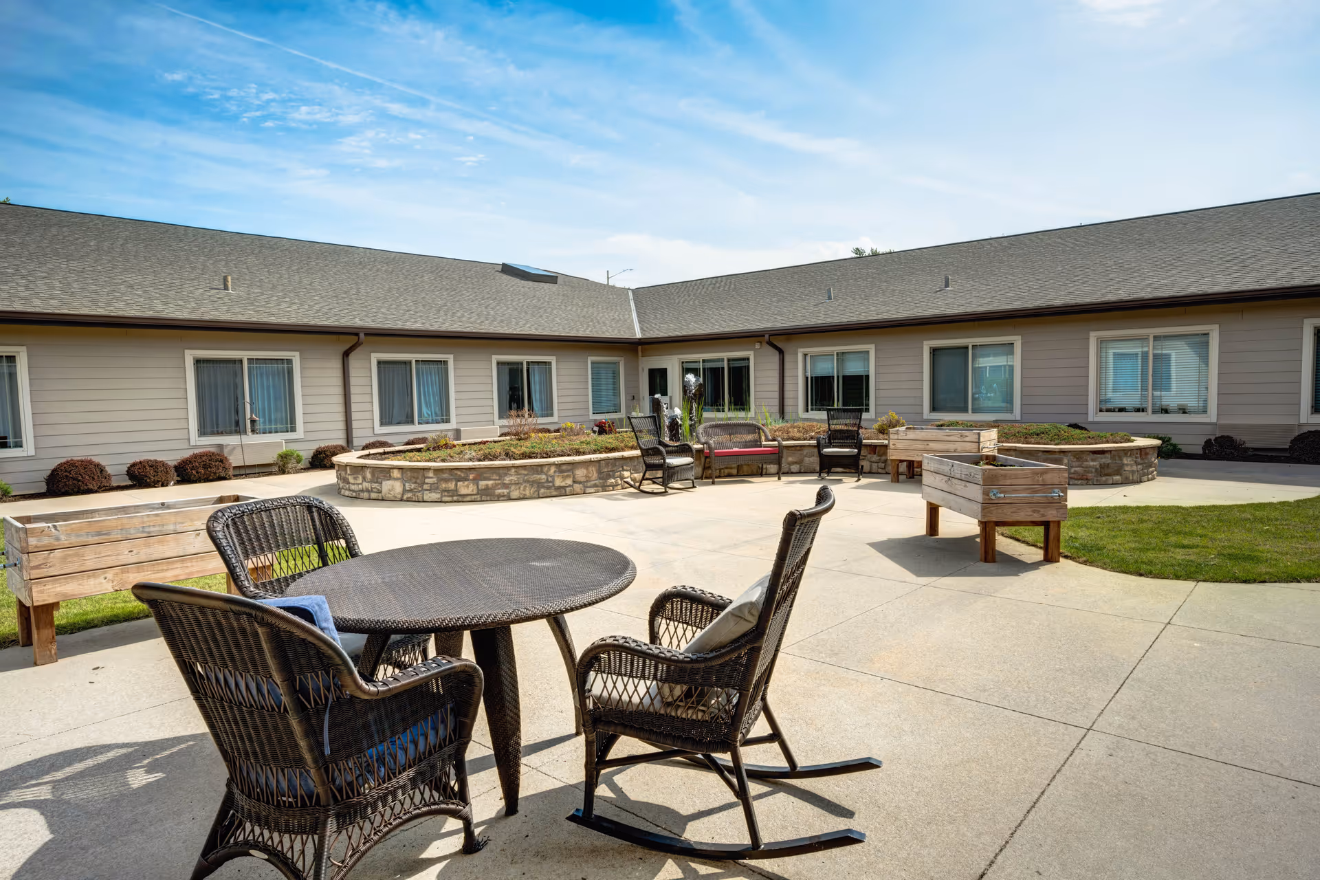 Outdoor courtyard area at Cedar Creek of Prairie Meadows featuring a round table with four wicker chairs, including a rocking chair, surrounded by raised garden beds and a stone planter with shrubs. The courtyard is enclosed by a single-story building with multiple windows under a clear blue sky.