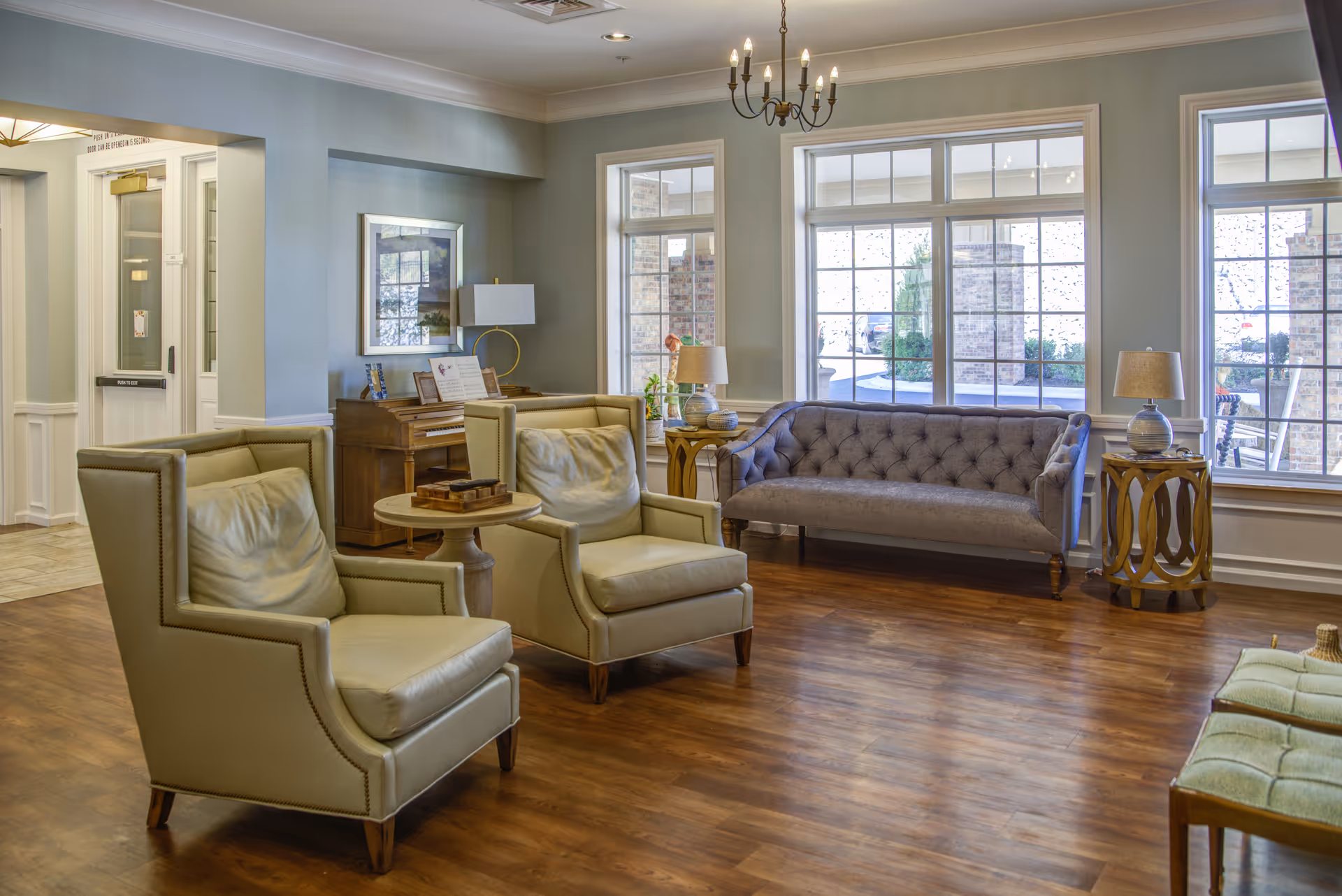A bright and spacious living room area in an assisted living facility featuring two beige armchairs with cushions, a tufted gray sofa, wooden side tables with lamps, large windows letting in natural light, a wooden piano against the wall, and a chandelier hanging from the ceiling.