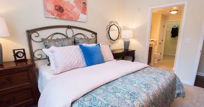 A neatly made bed with a decorative metal headboard, featuring patterned bedding in shades of blue, pink, and green, with multiple pillows including a prominent blue one. On the left side, there is a wooden nightstand with a lamp and a small clock. On the right side, there is a dark wooden desk with a lamp and a round decorative mirror on the wall above it. In the background, an open door reveals a bathroom with a toilet and a walk-in shower.