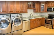 A kitchen area featuring wooden cabinets, a granite countertop, a stainless steel sink, a dishwasher, a front-loading washing machine and dryer, and a stainless steel oven with a microwave above it. The floor is light-colored wood.