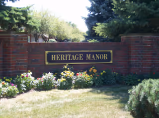 Brick entrance sign reading 'Heritage Manor' set in a landscaped lawn with flowers and trees.