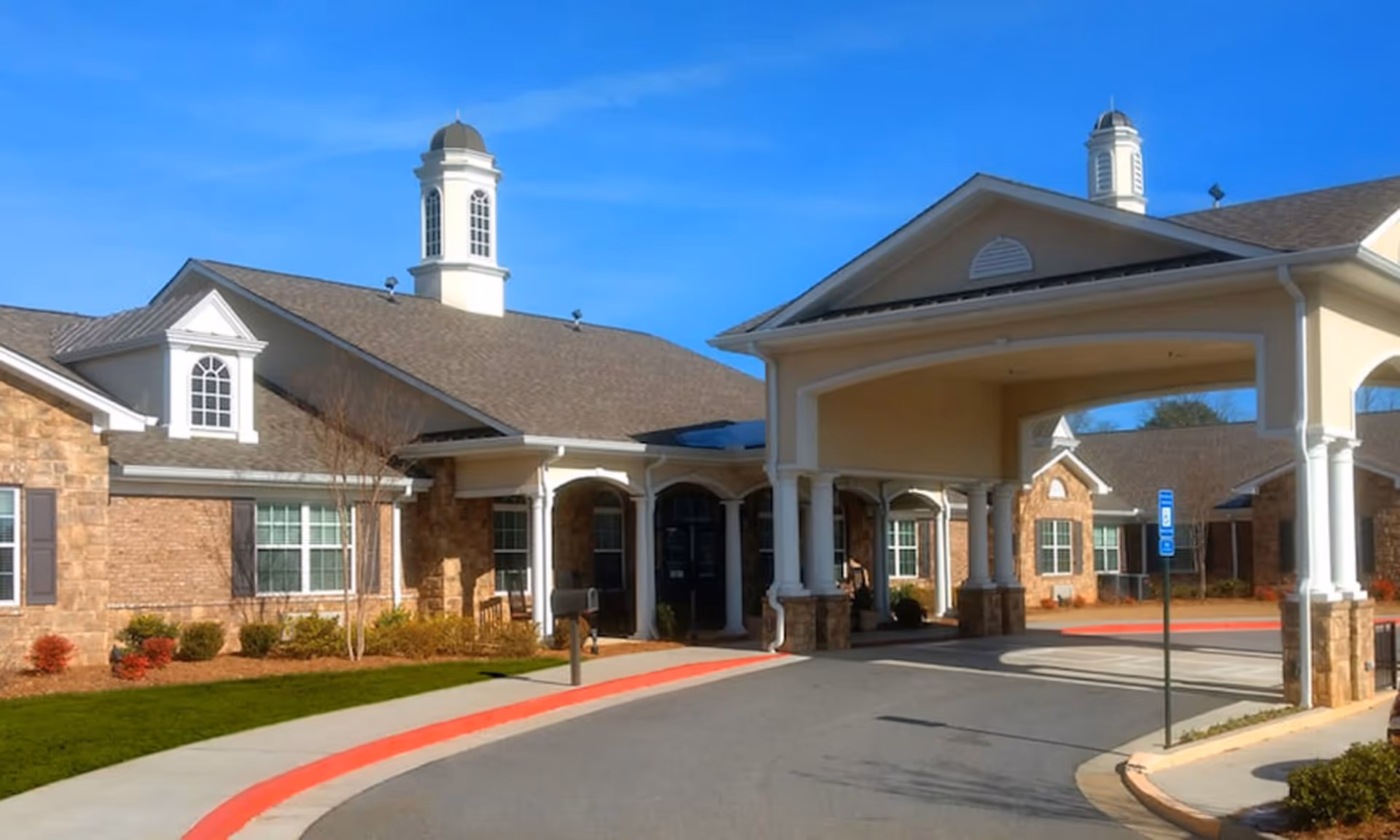 Exterior view of Addington Place Of Alpharetta senior living facility showing a large covered entrance with columns, brick and stone facade, multiple windows, and a clear blue sky.