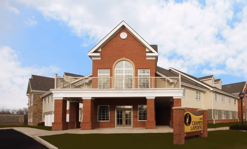 Front exterior of a two-story brick and siding senior living building with a covered entrance and a 'Cranes Landing' sign.