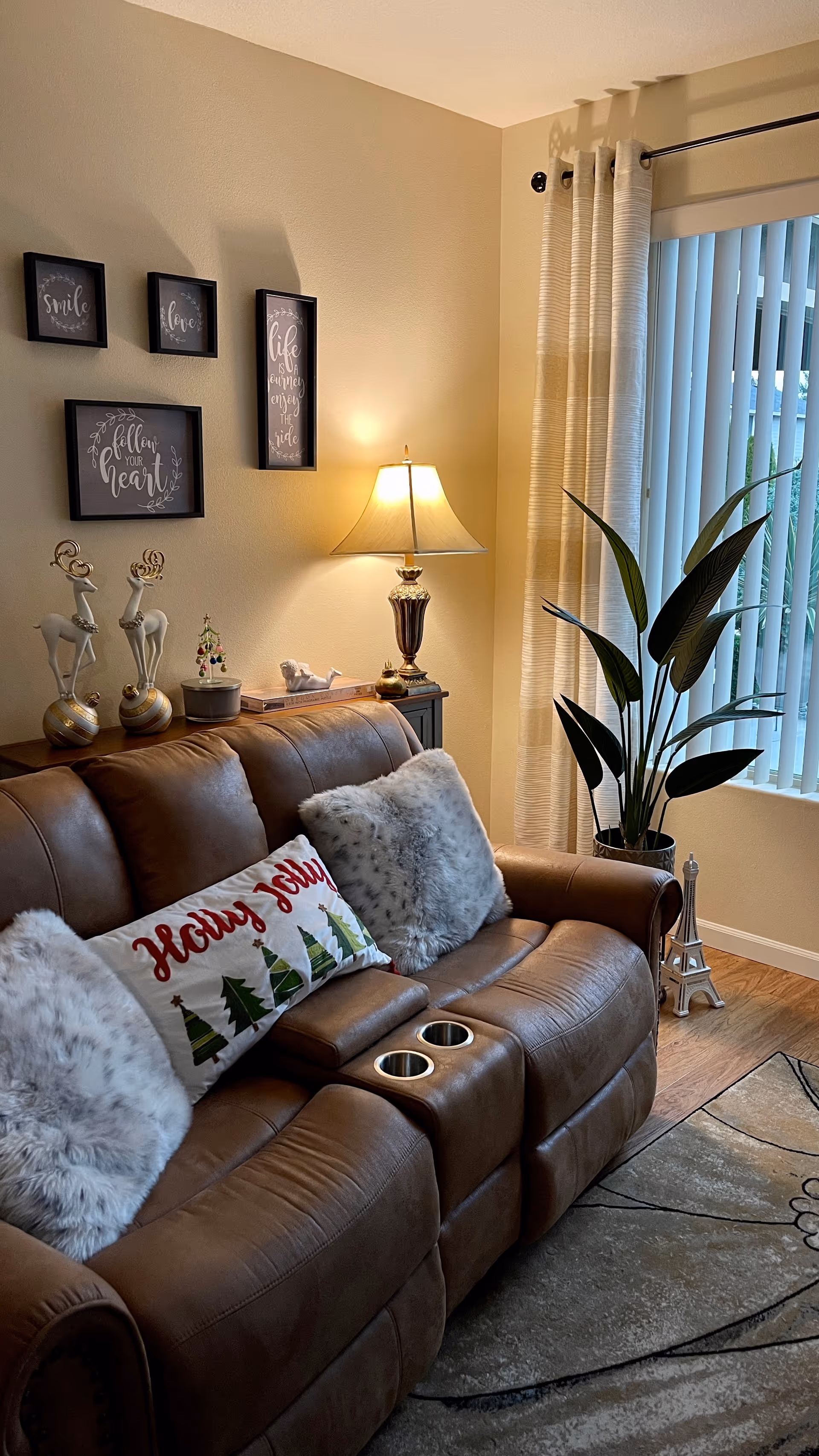 Cozy living room with a brown leather reclining sofa, decorative pillows and pillows, a side table lamp, wall art, and a potted plant by vertical blinds.