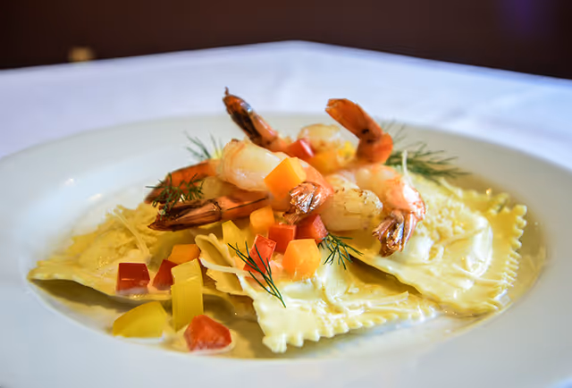 A white plate with ravioli pasta topped with cooked shrimp, diced red and yellow bell peppers, and garnished with fresh dill.