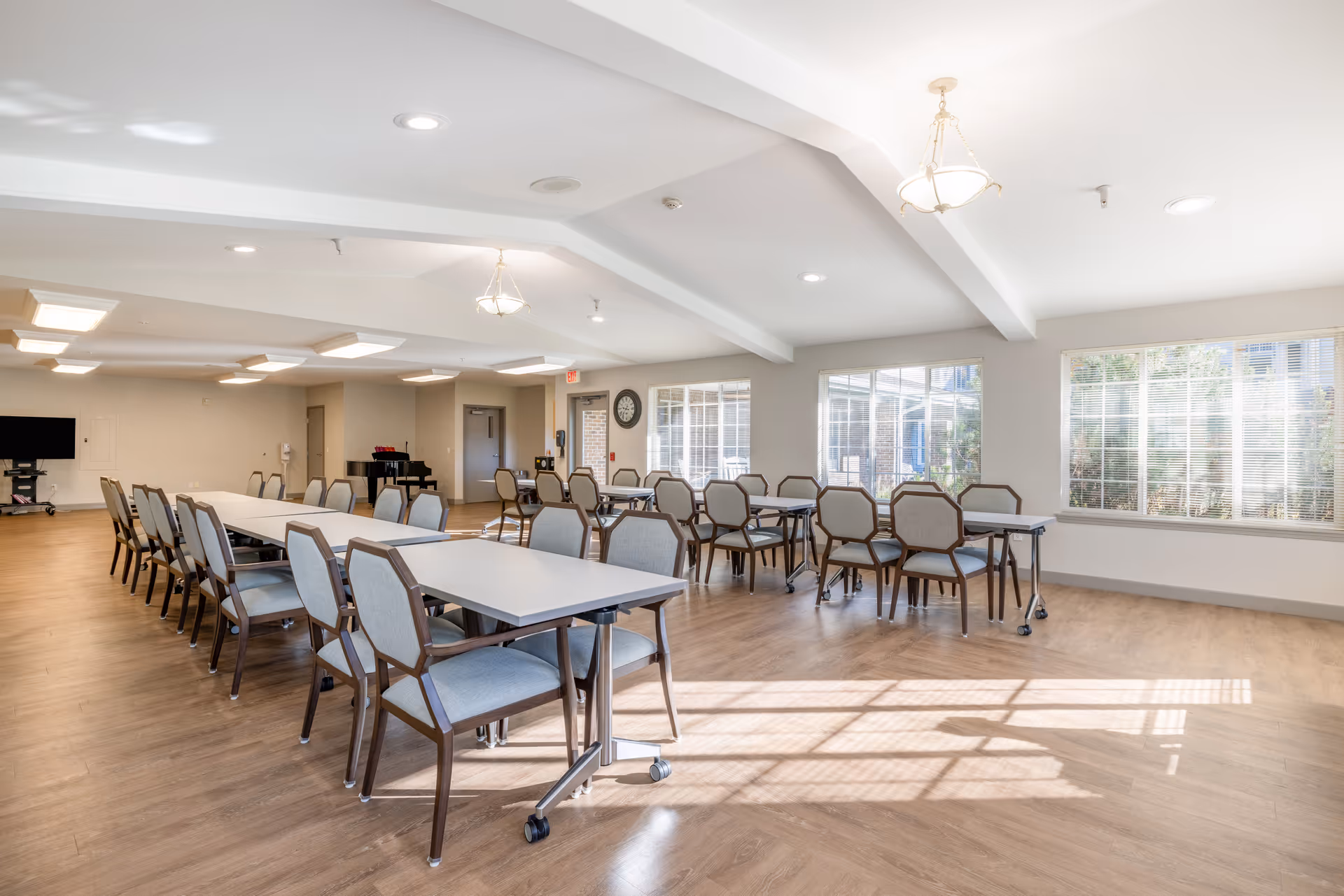 A bright and spacious dining room with multiple tables and chairs arranged neatly. Large windows on the right side allow natural light to fill the room. The floor is wood, and the ceiling has recessed lighting and hanging light fixtures. A clock is mounted on the wall near the windows, and a piano is visible in the background.