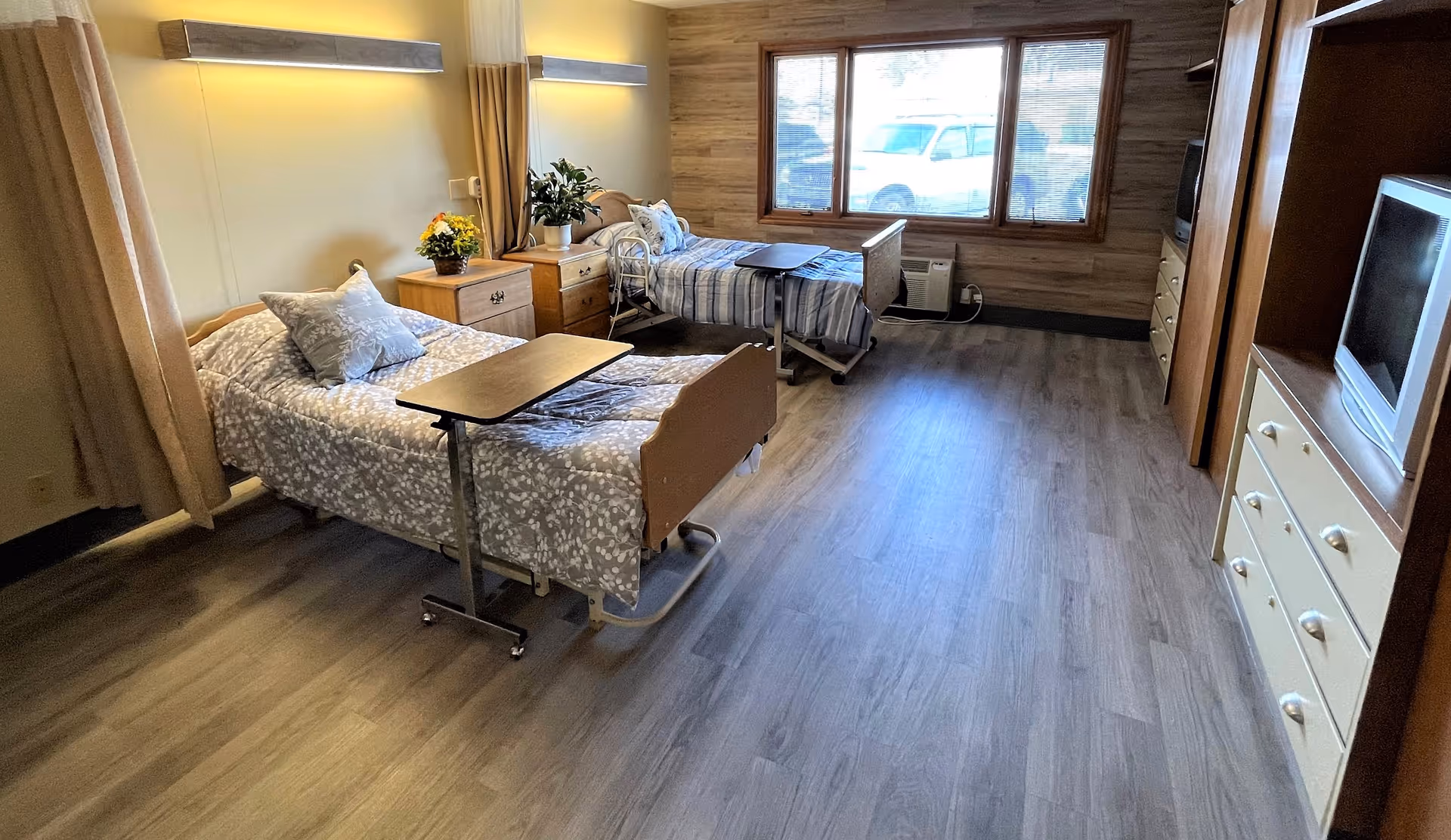 A well-lit room with two hospital-style beds, each with a small overbed table. The beds have patterned bedding and pillows. Between the beds is a wooden nightstand with a plant and flowers. The room has wood flooring, a large window with a view outside, and a dresser with a television on top. The walls are light-colored with wooden paneling on one side.