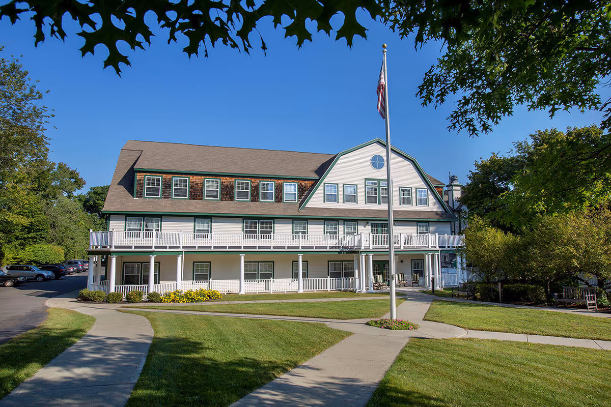Exterior view of a three-story senior living facility building with white siding and green trim, featuring multiple windows and a wrap-around porch. A flagpole with an American flag stands in front of the building, surrounded by well-maintained green lawns, sidewalks, and trees under a clear blue sky.