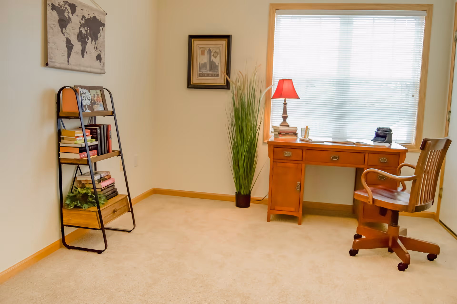 A bright room with beige carpet featuring a wooden desk with drawers, a red lamp, books, and a vintage typewriter on top. A wooden swivel chair is positioned in front of the desk. To the left, there is a black metal and wood bookshelf holding books and a small plant. A tall green plant is placed between the desk and the bookshelf. A large window with blinds allows natural light into the room. A framed picture and a world map hang on the walls.