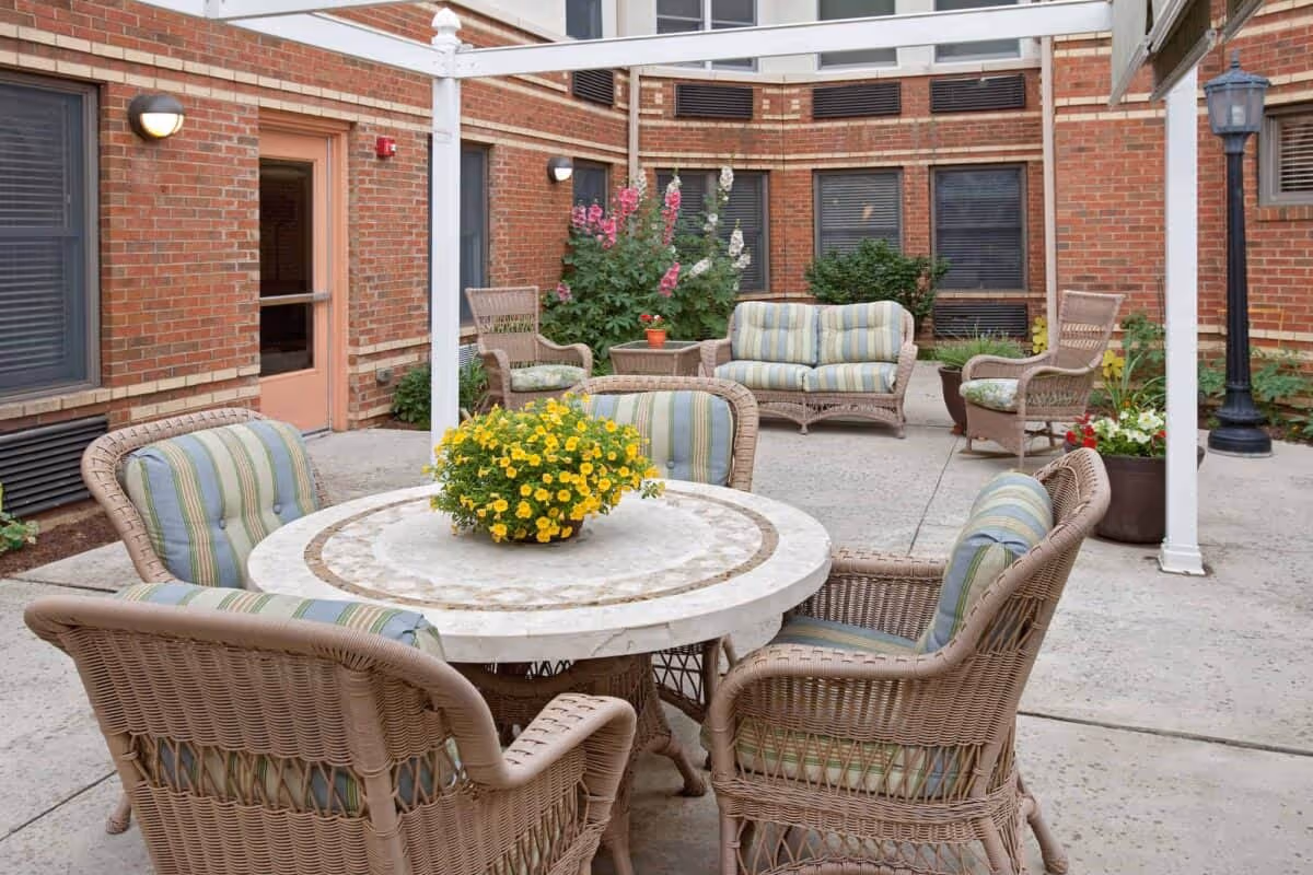 Outdoor patio area with wicker furniture including a round table with four cushioned chairs and a flower pot with yellow flowers on the table. In the background, there are additional wicker chairs and a loveseat with striped cushions, surrounded by brick walls and some plants.