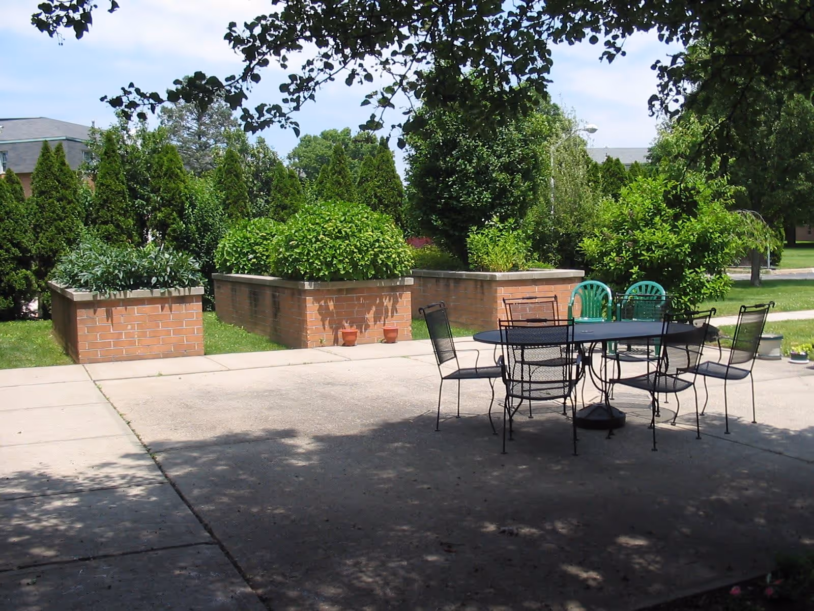 Outdoor patio area with a black metal table and six chairs, surrounded by green bushes and trees. There are three large brick planters filled with lush green plants in the background, and a clear blue sky above.