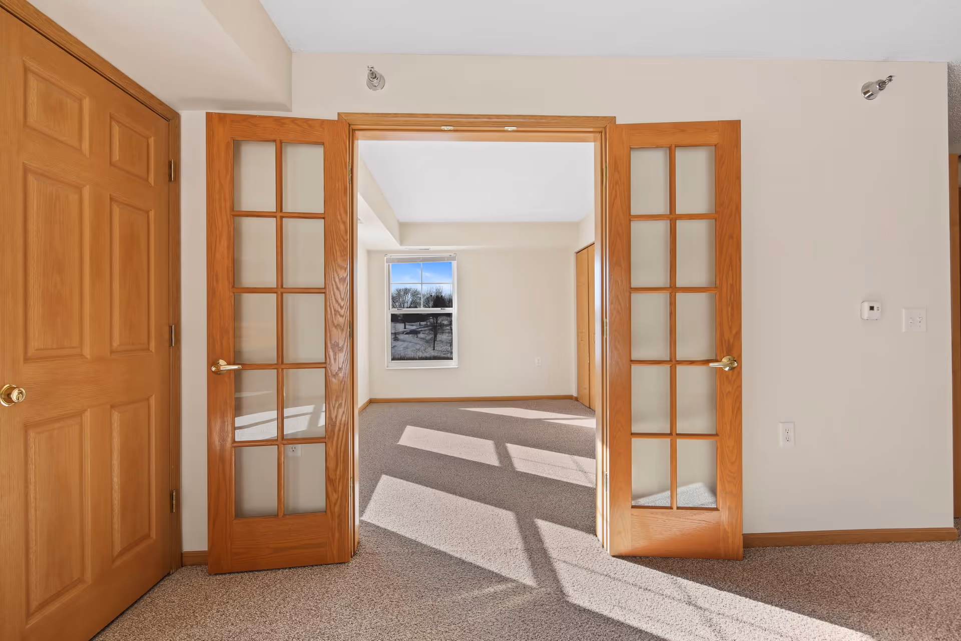 Empty room with beige walls and carpeted floor, featuring wooden double doors with glass panels open to another room with a window showing an outdoor view. There is a closed wooden door on the left and a thermostat on the right wall.
