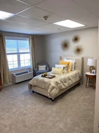A cozy bedroom with a single bed adorned with patterned bedding and multiple pillows. There is a small round wooden side table with a lamp and a framed photo next to the bed. A comfortable armchair with a pillow is placed near a large window with blinds and curtains. The walls are decorated with three sunburst-style wall hangings. The room has a carpeted floor and a ceiling with recessed lighting.