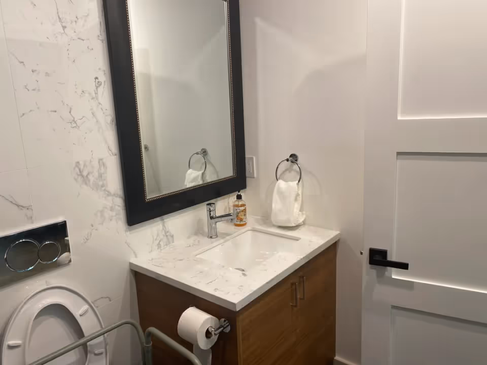 A modern bathroom featuring a white marble countertop with an under-mount sink, a chrome faucet, a soap dispenser, a towel ring with a white hand towel, a wall-mounted dual flush toilet, a toilet paper holder with a roll of toilet paper, a large rectangular mirror with a black frame, and a white paneled door with a black handle.
