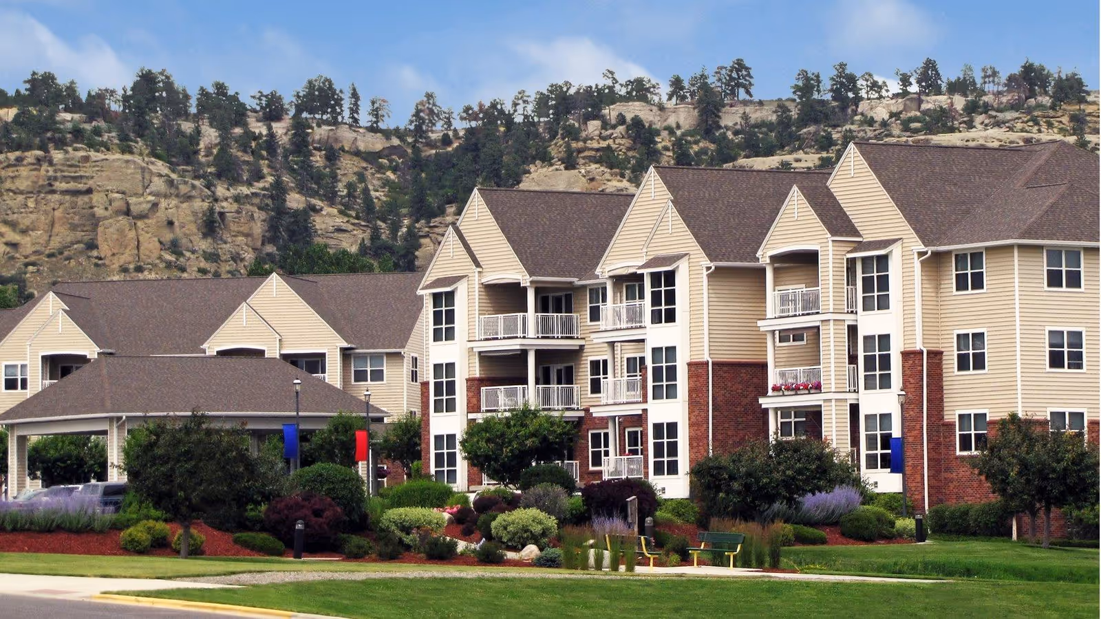 Exterior view of a multi-story senior living facility with beige siding and brick accents, surrounded by well-maintained landscaping including bushes, trees, and flower beds. There are benches and a covered entrance visible, with rocky hills and pine trees in the background under a partly cloudy sky.