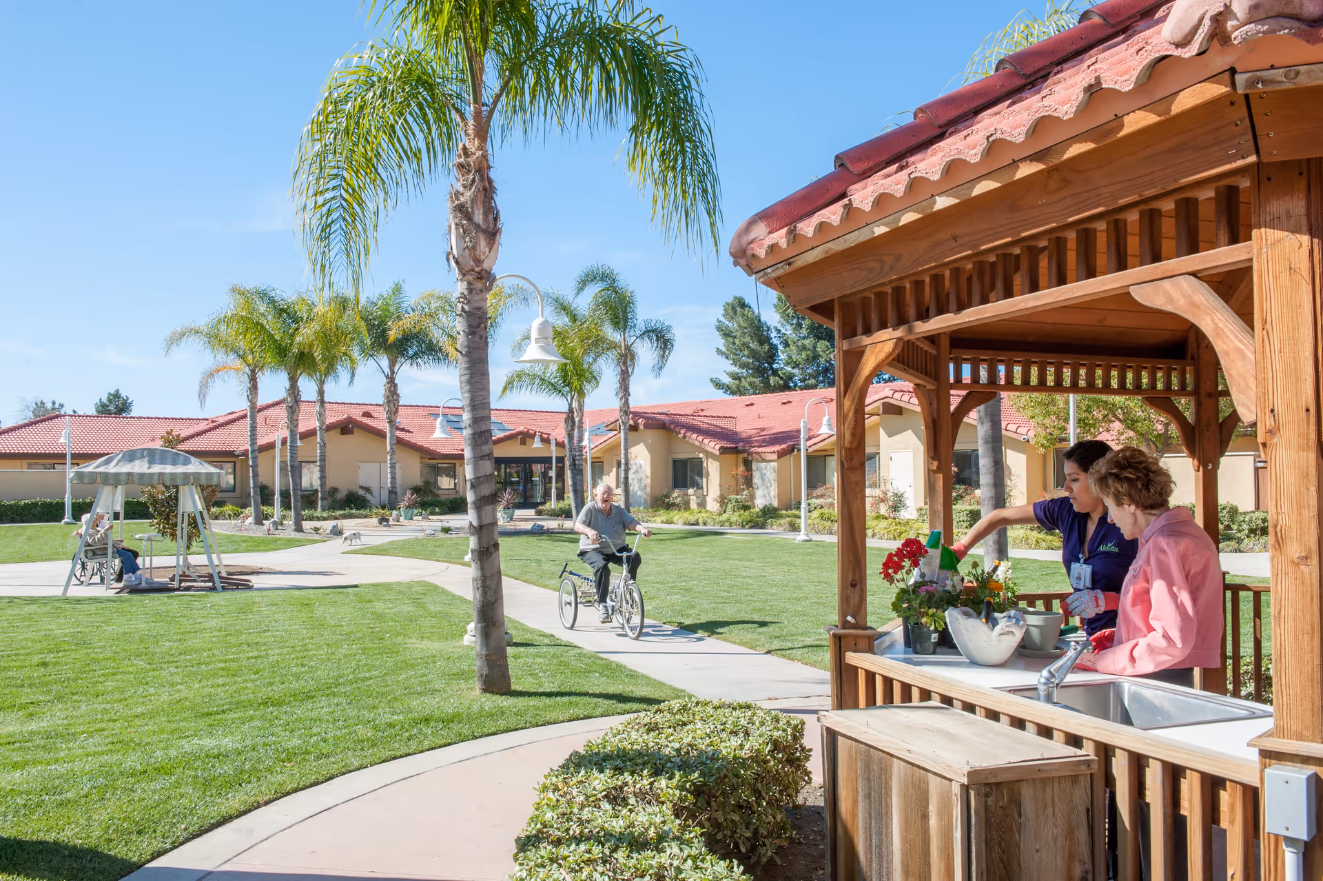 A sunny outdoor courtyard at Westmont of Riverside featuring a well-maintained lawn with palm trees, a paved walking path, and a wooden gazebo where two women are tending to potted plants. In the background, a man is riding a tricycle on the path, and another person is sitting on a swing under a canopy. The building with red-tiled roofs surrounds the courtyard.