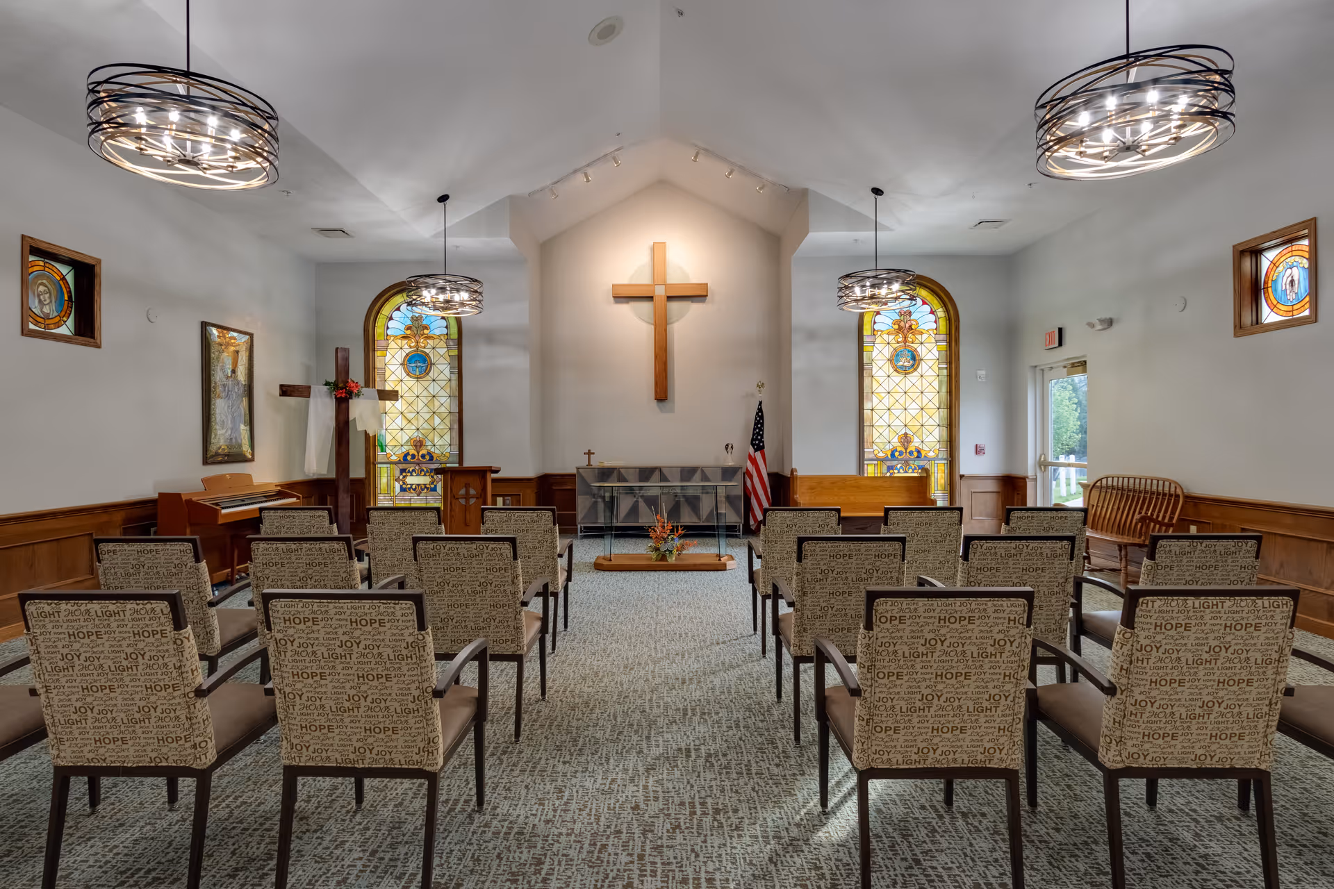 Interior view of a chapel with rows of chairs facing a wooden cross mounted on the front wall. The room features stained glass windows on either side of the cross, two modern circular chandeliers hanging from the ceiling, a piano on the left, and an American flag near the front. The walls are light-colored with wood paneling along the lower half.