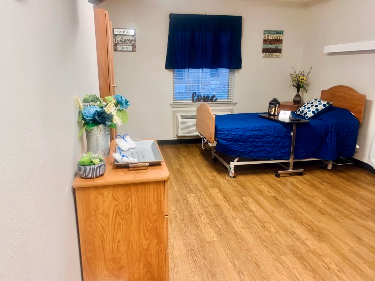 A neatly arranged bedroom in a nursing facility with a single bed covered in a blue quilt and a polka dot pillow. There is a small overbed table with a lantern and a tissue box on it. A wooden dresser with decorative plants and folded towels is positioned against the left wall. The room has wood flooring, a window with blinds and a dark blue valance, and motivational wall art.