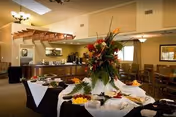 A spacious dining area with tables and chairs arranged around a buffet table covered with white and black tablecloths. The buffet table is decorated with a large floral centerpiece and plates of food. The room has warm lighting, a high ceiling with skylights, and a staircase leading to an upper level.