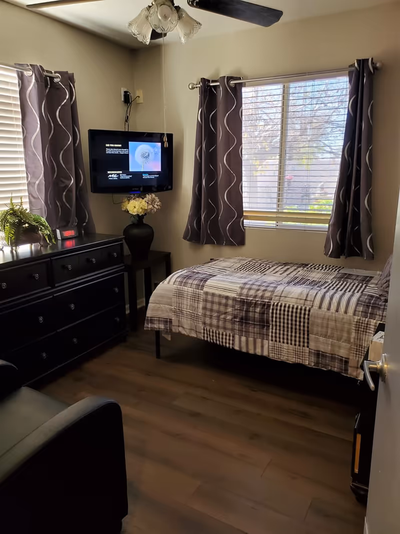 A small bedroom with a single bed covered in a patchwork quilt, a wall-mounted TV, a black dresser with plants and a digital clock on top, a ceiling fan with lights, and two windows with patterned curtains letting in natural light.