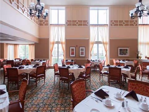 A spacious dining room with multiple round tables covered with white tablecloths, each set with cups, saucers, and folded black napkins. The room features large windows with sheer curtains allowing natural light to fill the space, patterned carpet, and elegant chandeliers hanging from the ceiling. Framed artwork is displayed on the walls.