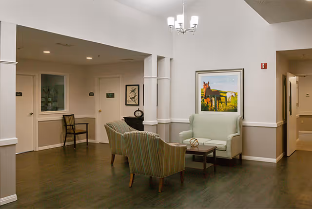 A seating area in a senior living facility with two striped armchairs and a light green loveseat around a small wooden coffee table. The walls are painted white and beige, with a framed picture of horses hanging above the loveseat. There is a chandelier light fixture on the ceiling and a wooden chair against the wall near two doors in the background.