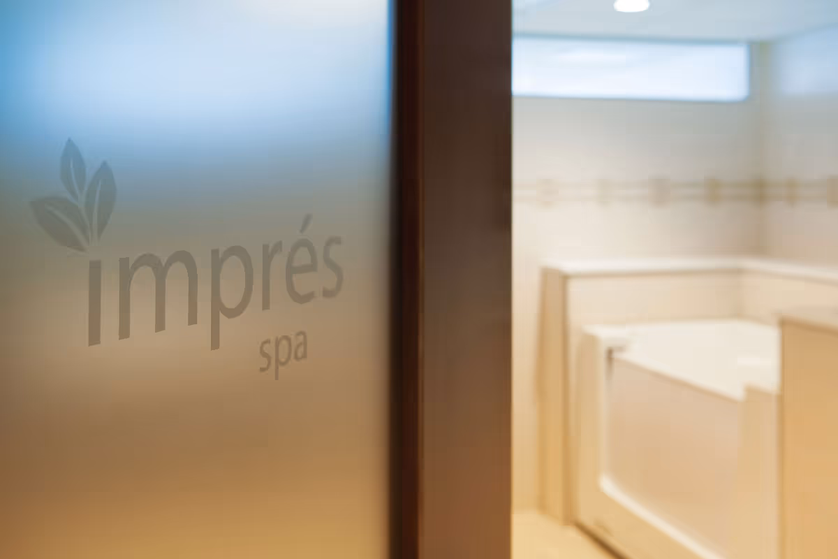 Frosted glass door with the words 'imprés spa' etched on it, partially open to reveal a clean, light-colored spa or bathroom area with a built-in bathtub and a small window near the ceiling.