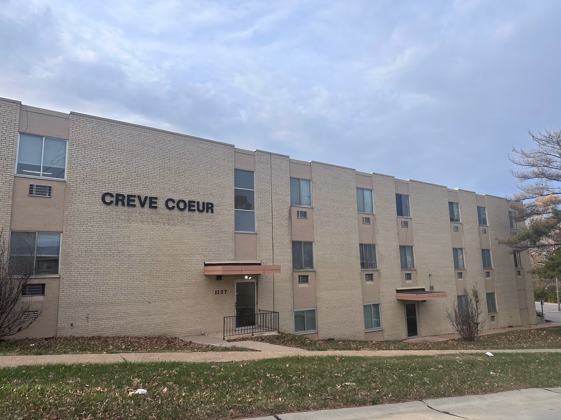 Exterior view of a three-story beige brick building with the name 'CREVE COEUR' displayed on the front. The building has multiple windows and two entrances with small awnings. There is a grassy area and a sidewalk in front of the building under a cloudy sky.