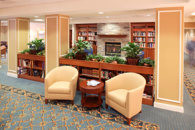A comfortable lounge area with two cream armchairs, a small round table, bookshelves, potted plants and a stone fireplace.