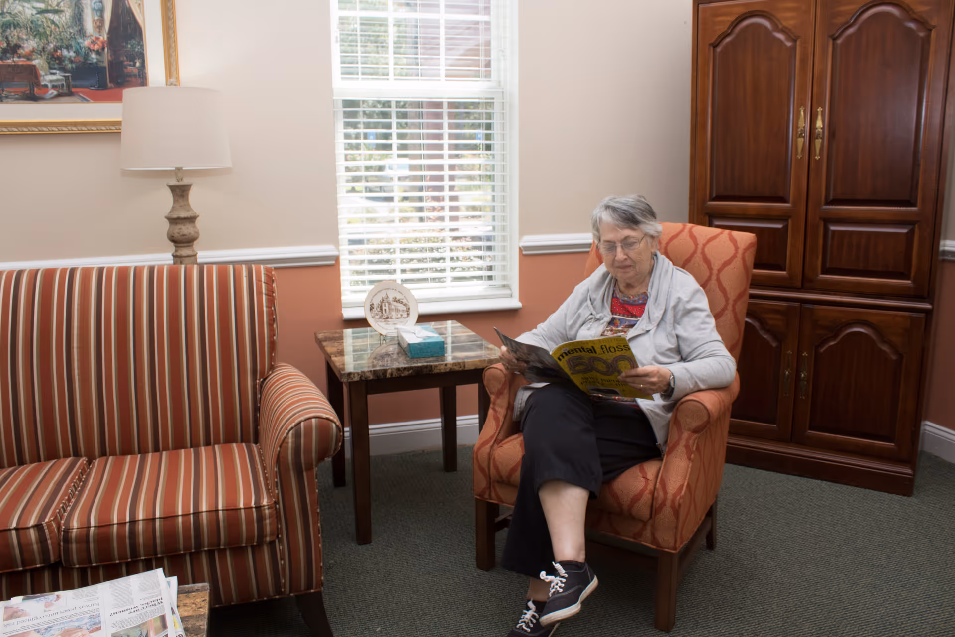 An elderly woman sits in an upholstered armchair reading a magazine in a cozy living room with a striped sofa, side table, lamp, and wooden cabinet.