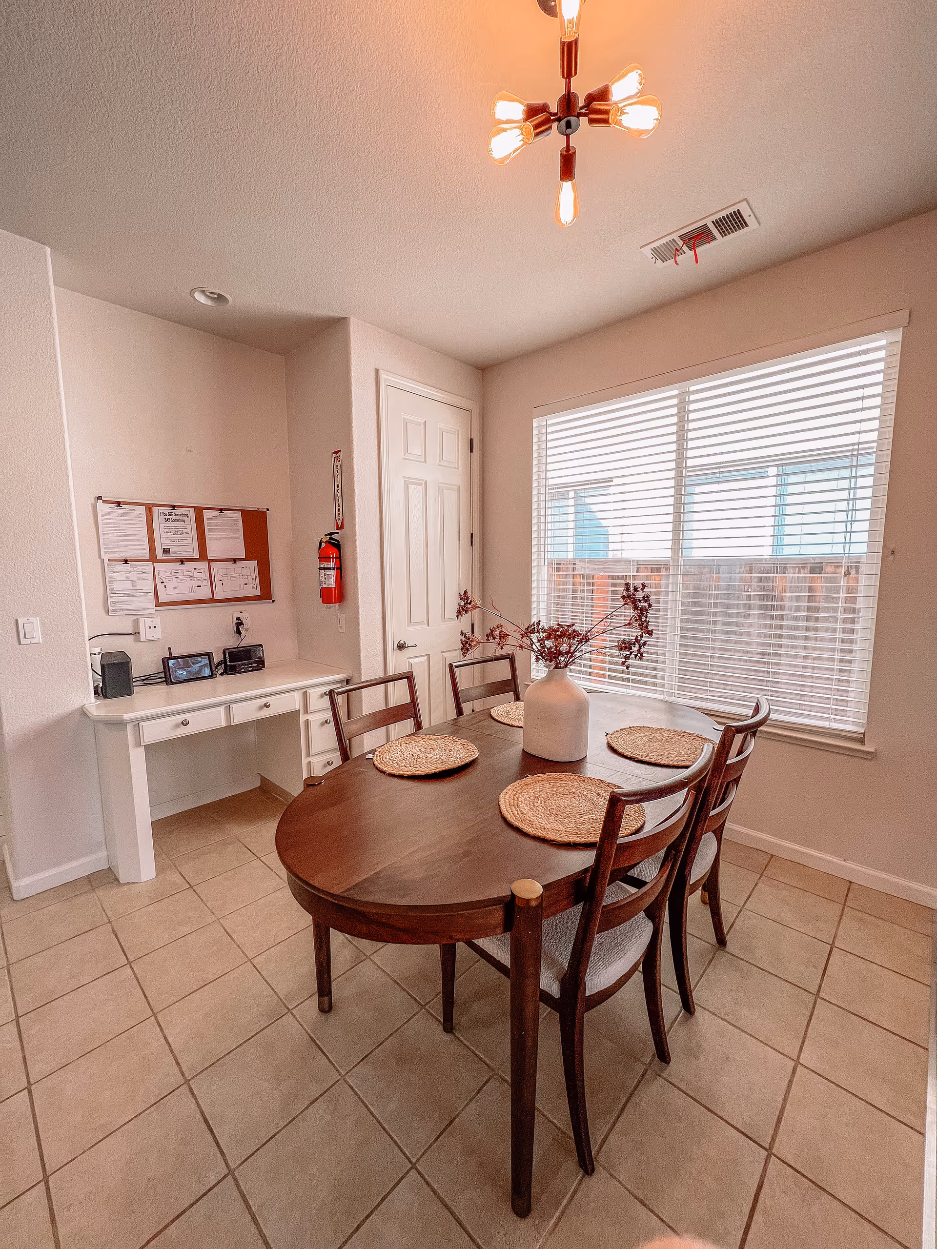 Dining area with a wooden table and four chairs topped with woven placemats and a vase, next to a large window and a small desk with a bulletin board.