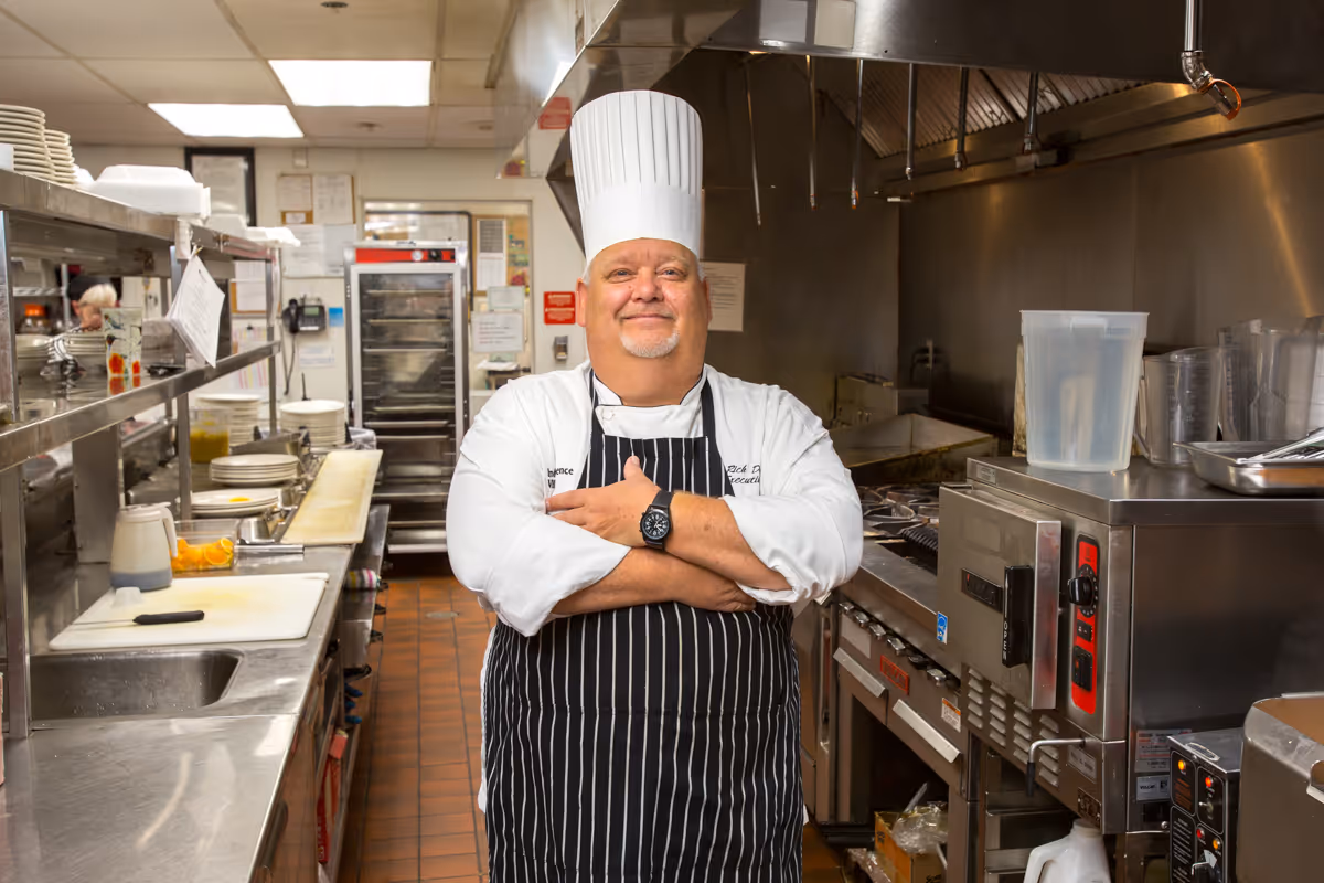 A chef wearing a white chef's hat, white coat, and black striped apron stands with arms crossed in a commercial kitchen. The kitchen has stainless steel counters, cooking equipment, and shelves with plates and kitchen tools.