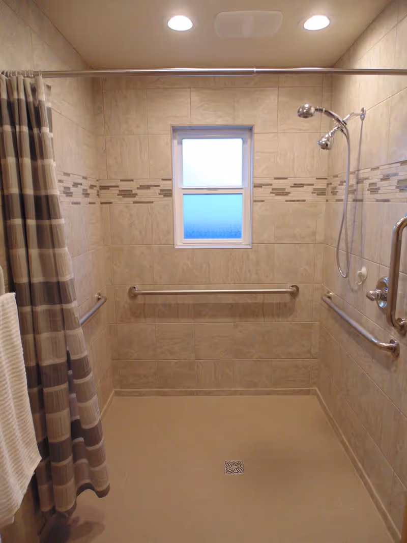 A tiled walk-in shower with beige and brown tiles, a window with frosted glass, a striped shower curtain, two shower heads, and multiple stainless steel grab bars for safety.