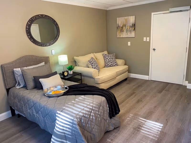 A cozy senior living bedroom featuring a single bed with gray bedding and multiple pillows, a small black side table with a lamp and a small plant, a beige loveseat with patterned cushions, a round decorative mirror on the wall, and a piece of abstract art above the loveseat. The room has light wood flooring and neutral-colored walls.