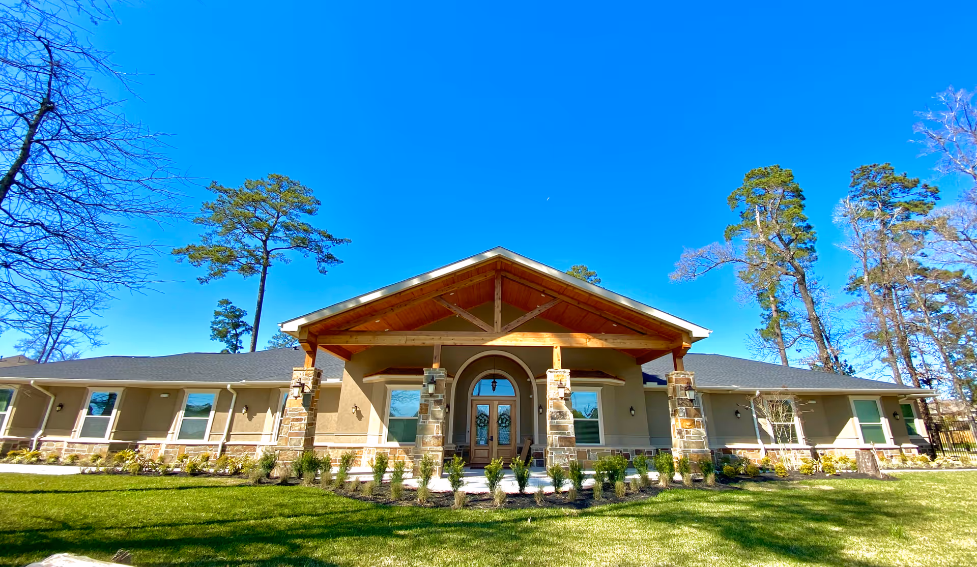 Single-story memory care community building with a covered wooden entrance supported by stone columns, a manicured lawn and tall trees under a clear blue sky.