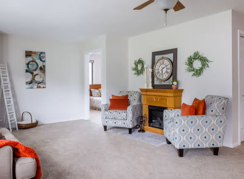 A cozy living room with two patterned armchairs featuring orange cushions, a wooden fireplace with a large clock and decorative wreaths above it, a beige carpet, and a ceiling fan. A doorway leads to another room with a bed visible.