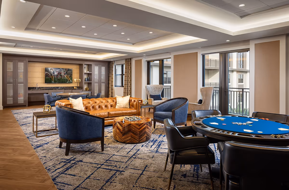 A spacious and well-lit senior living common area featuring a brown leather tufted sofa, blue upholstered chairs, a round wooden coffee table, and a poker table with cards and chips. The room has large windows with views of the building exterior, a patterned carpet, and a ceiling with recessed lighting. In the background, there is a wall-mounted TV showing a football game and built-in cabinetry with glass doors.