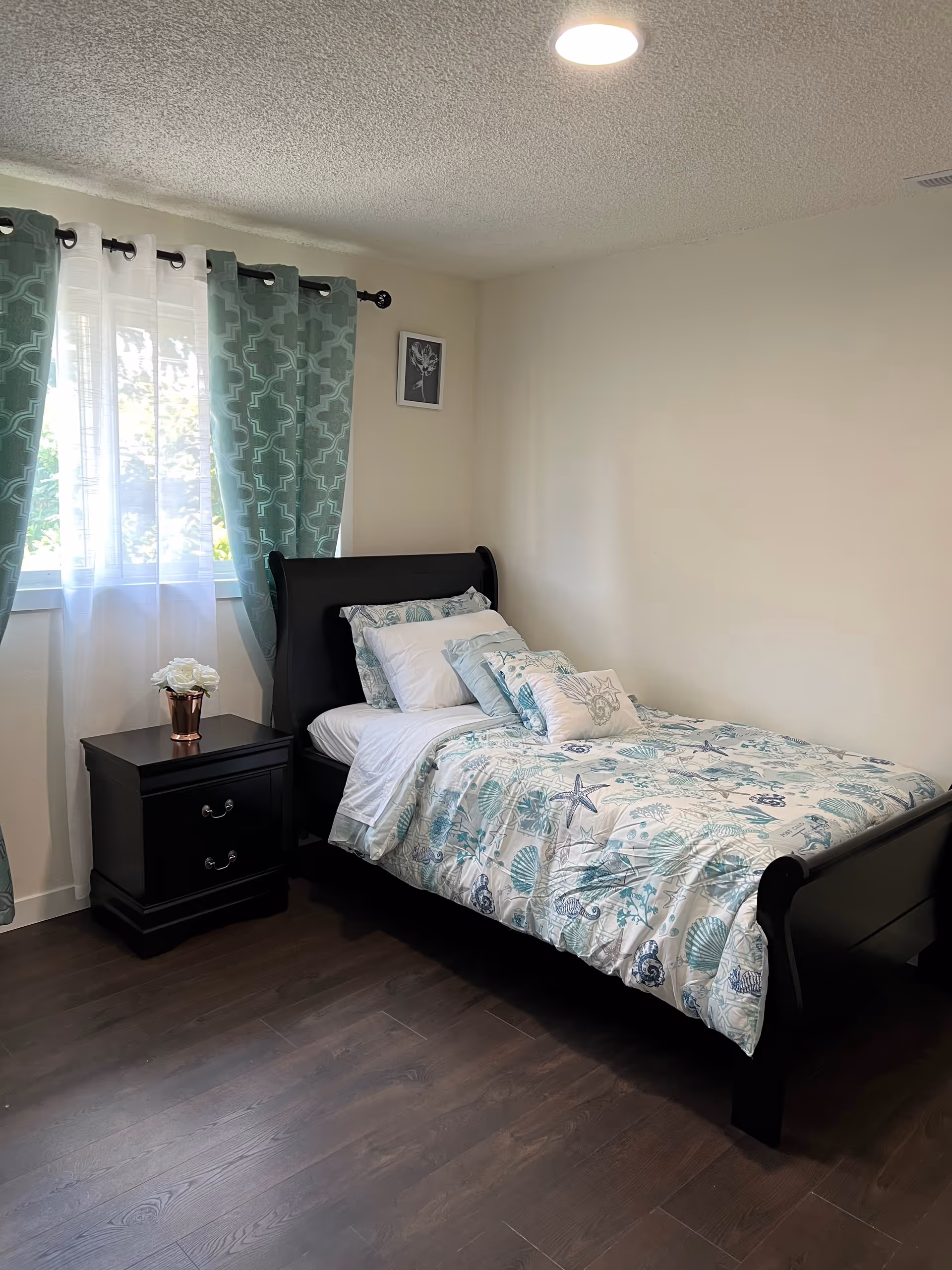A neatly made single bed with a dark wooden frame and sea-themed bedding in a bright room. Next to the bed is a dark wooden nightstand with two drawers and a small vase of white flowers. The room has light-colored walls, a window with sheer white curtains and teal patterned drapes, and dark wood flooring.