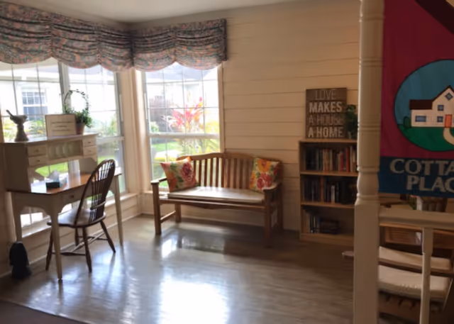 A cozy interior corner of a room with a wooden bench adorned with two floral cushions, a small wooden desk with a chair near large windows letting in natural light, a bookshelf with books and a decorative sign that reads 'LOVE MAKES A HOUSE A HOME', and a colorful banner with a house illustration partially visible on the right.