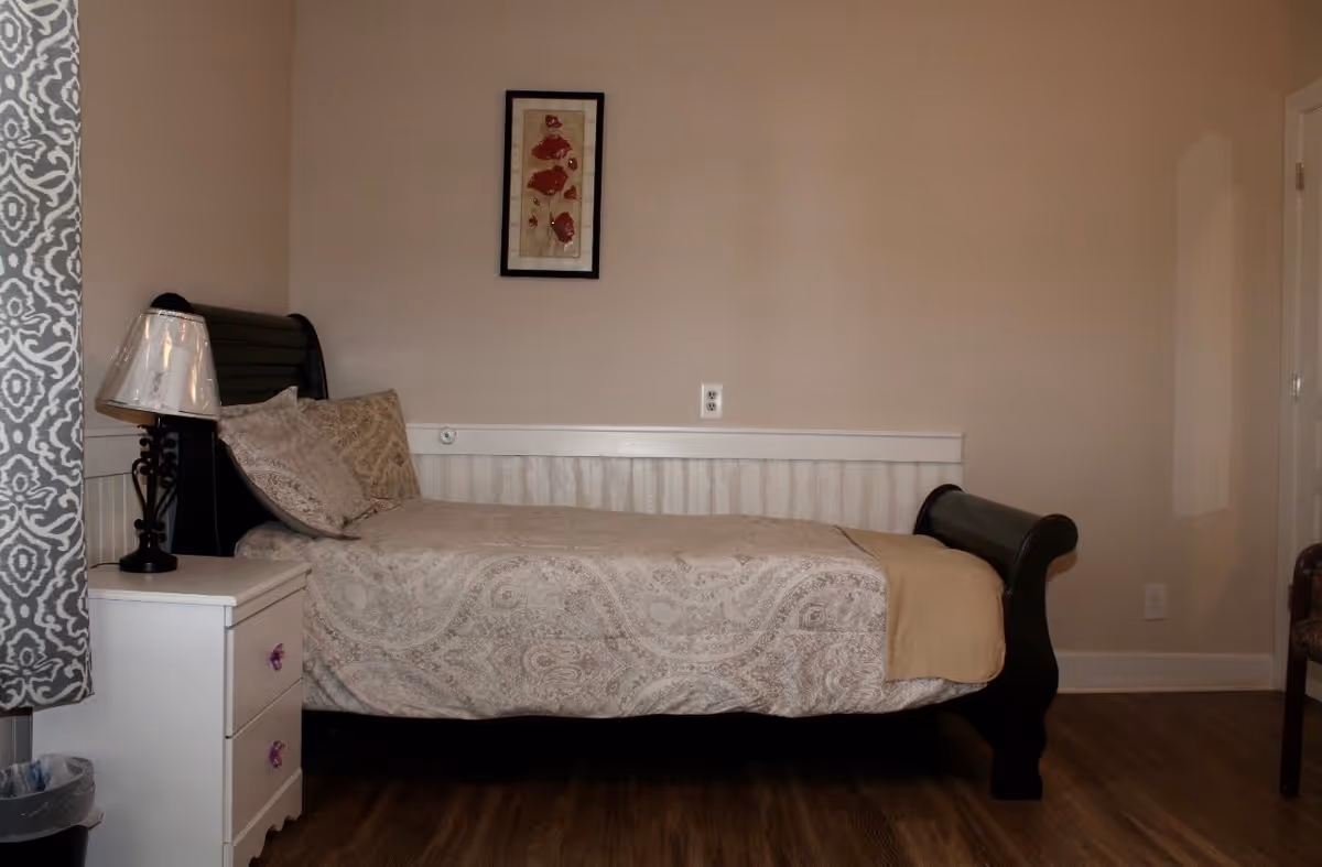 A single bed with a dark wooden frame and beige patterned bedding is positioned against a beige wall. Next to the bed is a white nightstand with two drawers and a lamp with a transparent shade. A framed artwork featuring red flowers hangs above the bed. The floor is wooden, and a patterned curtain is partially visible on the left side.