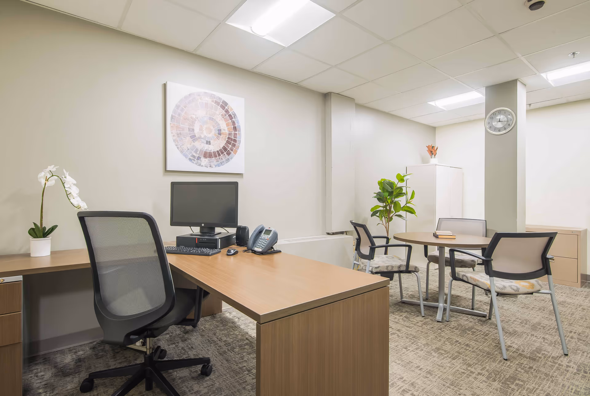 A clean and well-lit office space with a wooden desk, a black mesh office chair, a computer monitor, keyboard, mouse, and a telephone on the desk. There is a round meeting table with four chairs in the background, a potted plant, a white cabinet, and a wall clock mounted on a pillar. A decorative circular artwork hangs on the wall above the desk.